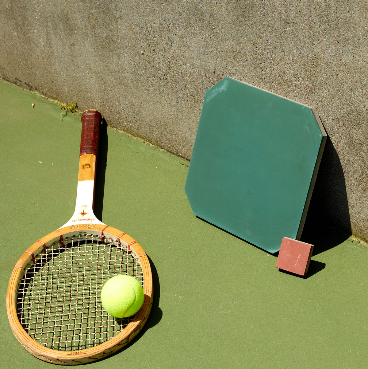 a tennis racket and ball along with a green cement tile displayed on a tennis court next to a cement wall